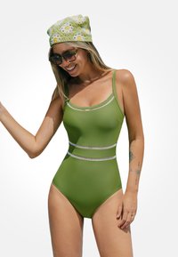 Bañador verde de una sola pieza con tirantes finos, que presenta una banda de acento de rayas horizontales. El modelo lleva un sombrero verde estampado y gafas de sol.