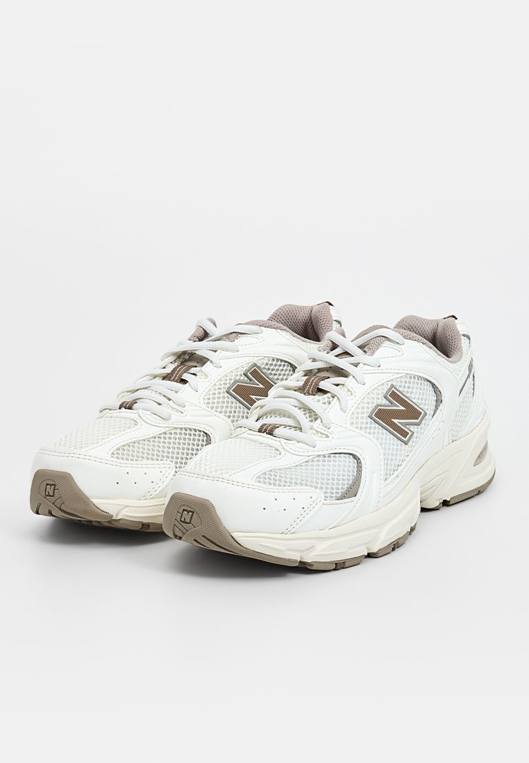 Zapatillas deportivas blancas con materiales de malla y sintéticos. Las características incluyen una suela de goma, acentos texturizados y un logo "N" en color marrón.