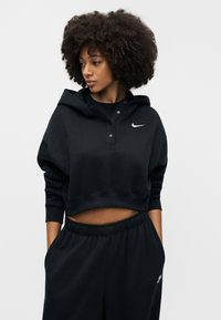 Schwarzer kurz geschnittener Hoodie aus weichem Material mit einer Fronttasche, Knopfleiste und einem kleinen weißen Nike-Swoosh-Logo auf der Brust.