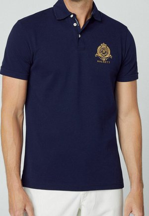 Tummansininen polo-paita, joka on valmistettu puuvillasta. Paita on kauluspaitamalli, jossa on kolme nappia ja kultaiset brodeeratut logot vasemmassa rinnassa. Lyhyet hihat.