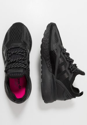 Paire de chaussures de sport noires avec semelles texturées et lacets à motifs, l'une montre une semelle intérieure rose vif avec un logo.