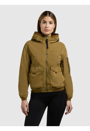 Donna che indossa una giacca marrone con cappuccio, tasche frontali e cerniera, mostrata dalla vita in su su uno sfondo semplice.