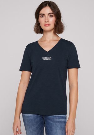 Woman with short wavy brown hair wearing a dark blue V-neck T-shirt with "SOCCX TIMELESS EDITION" text and blue jeans, standing against plain background.