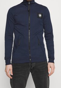 Marinblå zip-upp sweatshirt med hög krage, två sidofickor, ribbade ärmslut och nederkant, med en svart logotypapplikation på vänster bröst.