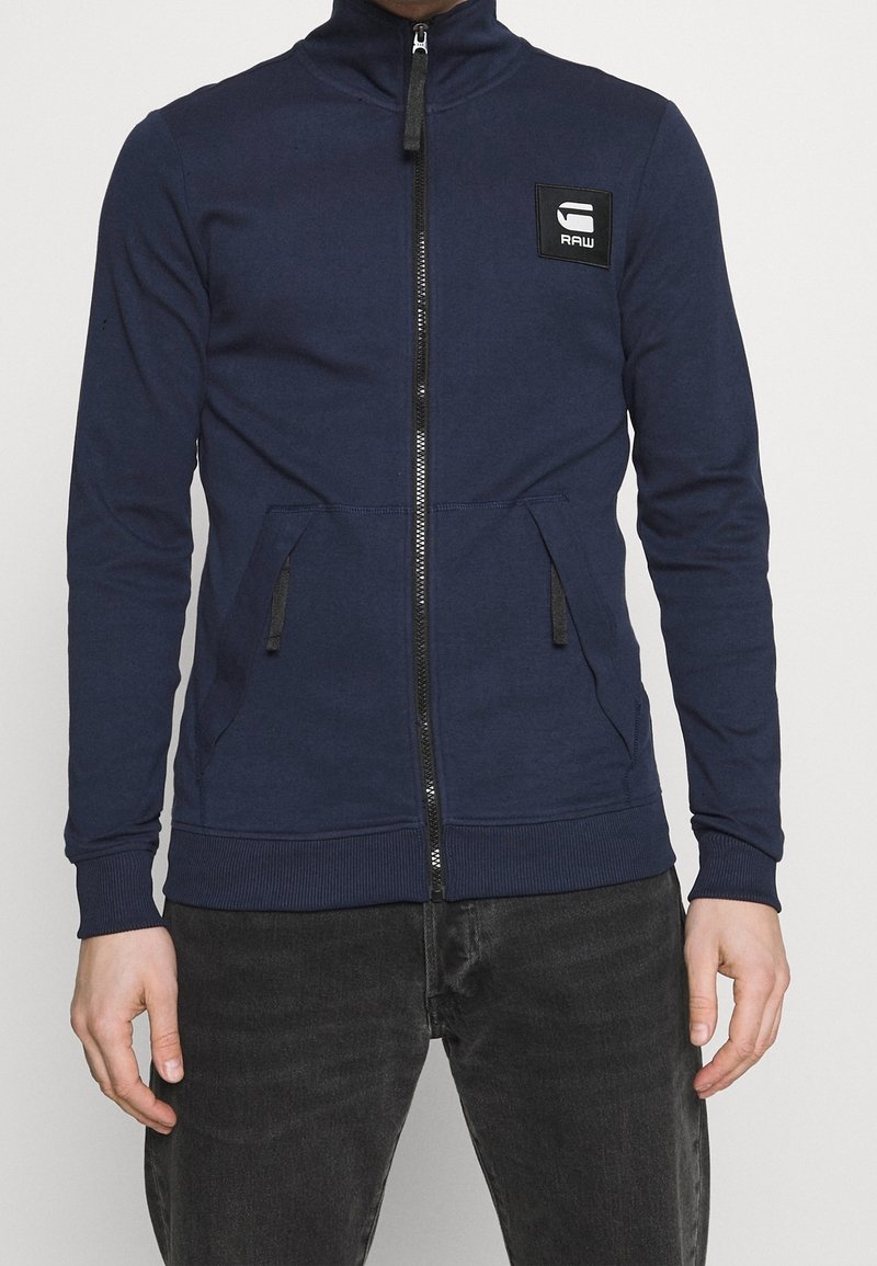 Marinblå zip-upp sweatshirt med hög krage, två sidofickor, ribbade ärmslut och nederkant, med en svart logotypapplikation på vänster bröst.