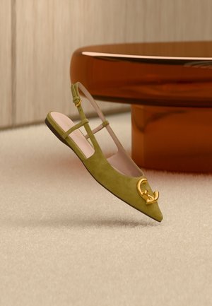 Oliiviroheline seemisnahast slingback-tasaotsaga king, millel on terav nina ja kuldse topelttähisega G-logoga ornament, eksponeeritud beežil vaibal pruunist ümmarguse lauapea kõrval.