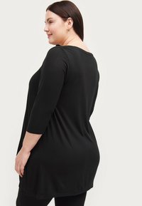 Sort tunika top med 3/4 ærmer, blødt stof, buet kant og en afslappet pasform. Bagfra ses den rundede halsudskæring og glatte tekstur.