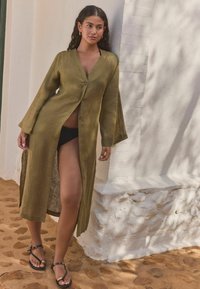 Olivově zelený lněný dlouhý cardigan s hlubokým výstřihem do V, zapínáním na knoflíky a širokými rukávy. Spárováno s černým plavkami a plochými sandály.