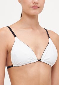 Hvit triangulær bikinitopp med svarte stropper og sømmer, med en mellom cutout-detalj og glatt, matt tekstur.