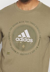 Olivengrøn bomulds-t-shirt med et cirkulært grafisk design featuring "adidas" logo i hvid, tre striber og tekstaccent. Korte ærmer.