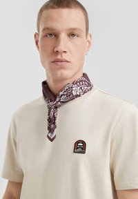 T-shirt in cotone beige con scollatura rotonda, caratterizzata da una spilla marrone e nera. Una bandana bordeaux scuro con motivi floreali bianchi è annodata intorno al collo.