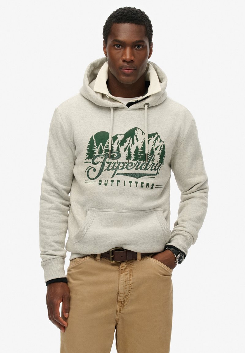 Felpa grigia con cappuccio, dotata di tasca a marsupio frontale, con una grafica verde di montagne e alberi. Indossata con pantaloni beige e una cintura.