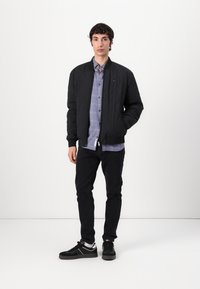 Black quilted bomber jacket with elastic cuffs, layered over a light blue plaid shirt, paired with black pants and black sneakers.