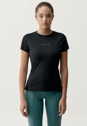 Jeune femme debout, portant un T-shirt noir ajusté avec un petit logo blanc et des leggings taille haute turquoise, fond neutre.