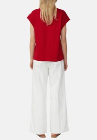 Vrouw met lang blond haar, gekleed in een rood T-shirt met korte mouwen, witte wijde broek en bruine sandalen, staande met de rug naar de camera op een witte achtergrond.