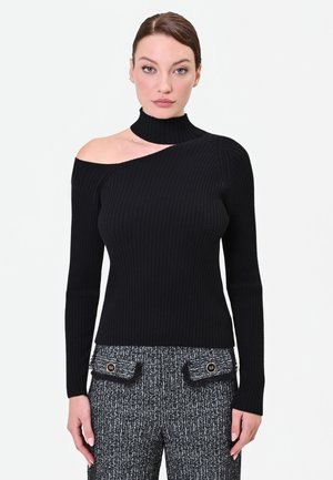 Donna che indossa un top nero a coste con maniche lunghe e una spalla con ritaglio asimmetrico, e pantaloni grigi con motivo e tasche frontali.