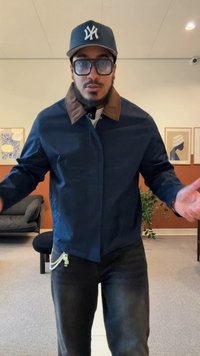 Chaqueta azul marino con cuello marrón, cierre de cremallera y bolsillos laterales. Lleva puestos jeans de mezclilla oscuros. Gafas grandes y transparentes.