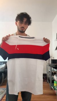 Young man holding up a white t-shirt with red and navy blue horizontal stripes in a living room with wooden floor.