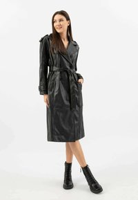 Cappotto trench in pelle nera con design doppiopetto, vita cinturata, dettagli sulle spalle e tasche laterali. Indossato con stivali neri stringati.