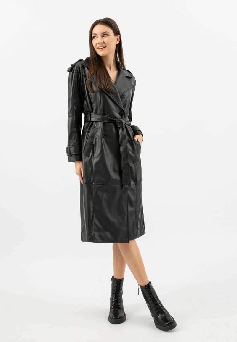 Cappotto trench in pelle nera con design doppiopetto, vita cinturata, dettagli sulle spalle e tasche laterali. Indossato con stivali neri stringati.