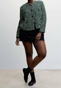 Veste texturée verte et blanche avec un col arrondi et des boutons noirs, associée à une mini-jupe noire et des bottines.