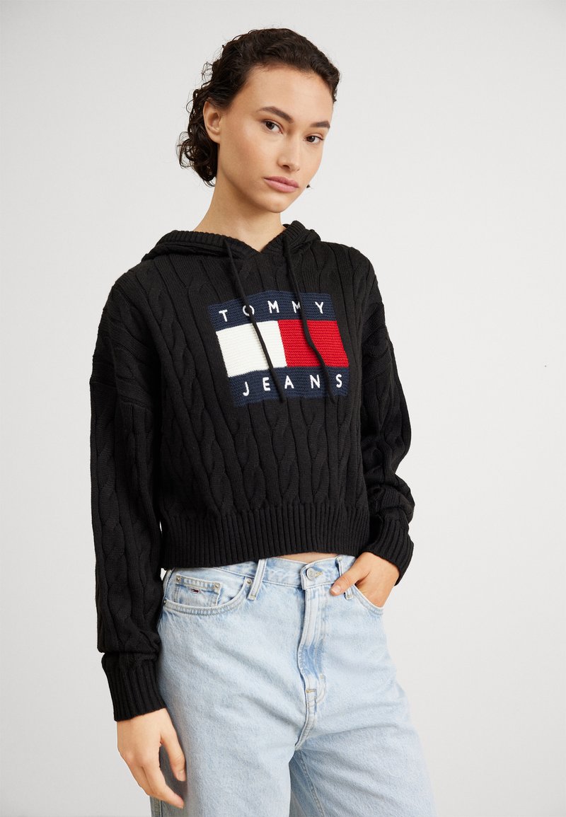 Kvačkana crna kapuljača s logom na prednjem dijelu koji prikazuje crvene, bijele i plave pravokutnike. Rebrasti manžeti i rub, nosi se s svijetlim trapericama.