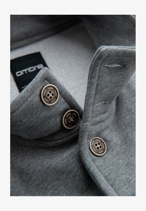Grå kravebeklædning med tekstureret stof, der har to bronzefarvede knapper og et sort mærke med "ombre" branding.