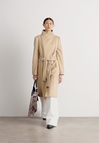 Abrigo clásico beige con un cuello alto y cinturón, combinado con pantalones anchos blancos. Bolsa negra y bufanda estampada en la mano, fondo simple.