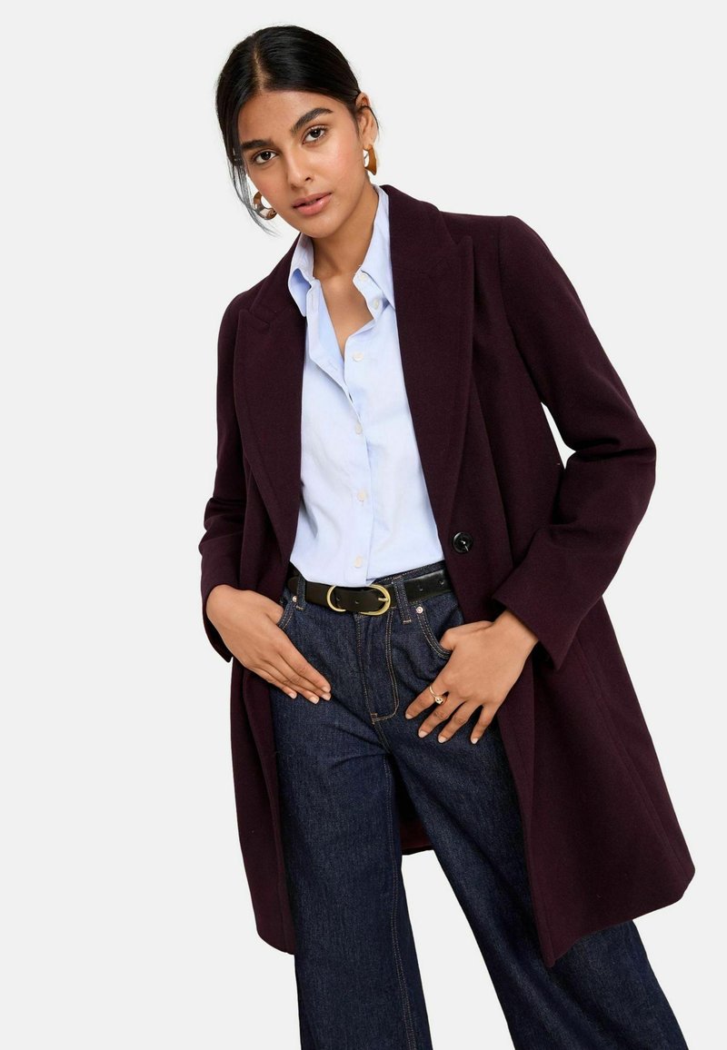 Cappotto bordeaux con un design sartoriale, caratterizzato da un bottone singolo, ab binato a una camicia azzurra chiara e pantaloni di denim scuro. Una cintura nera accentua l'outfit.