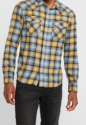 Homme portant une chemise à carreaux jaune, bleue et noire avec deux poches avant, associée à un pantalon noir, debout devant un fond blanc.