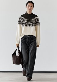 Femme marchant, portant un pull à motifs noir et blanc, un jean noir ample, des chaussures marron, et tenant un sac à main en daim marron foncé.