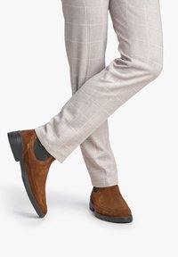 Bottines en suédine marron avec des inserts élastiques sur les côtés, associées à un pantalon à carreaux gris clair. Le pantalon a une coupe slim et une texture lisse.
