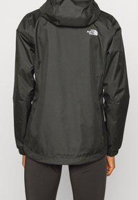 Svart vattentät jacka med huva, med en logotyp på övre ryggen, långa ärmar och en rak skärning för enkel rörelse.