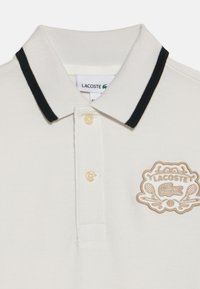 Бяла поло риза Lacoste с черна райета яка, два бутона отпред и бродиран лого с тенис ракети и крокодил.