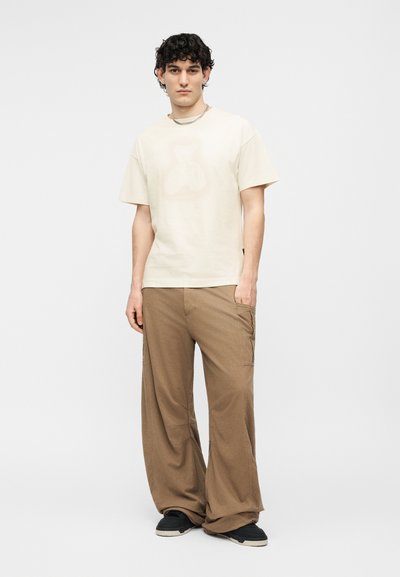 Νεαρός άνδρας που στέκεται, φοράει μπεζ κοντομάνικο πουκάμισο, φαρδιά καφέ παντελόνια, μαύρα αθλητικά παπούτσια και ασημένιο αλυσίδα λαιμού σε λευκό φόντο.