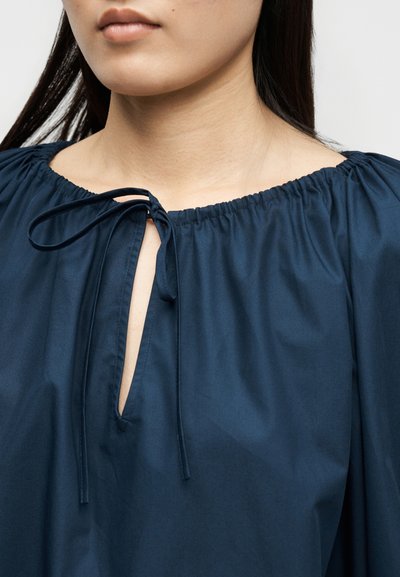 Krk a ramená ženy v námornícky modrej blúzke s elastickým výstrihom a zaväzovacím otvorom na kľúčik vpredu.