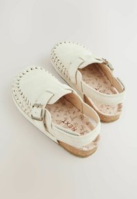 Sabots en daim blanc avec un bord tressé, une boucle réglable et une semelle en liège. Semelle intérieure à motif floral, design ouvert à l'arrière pour un enfilage facile.