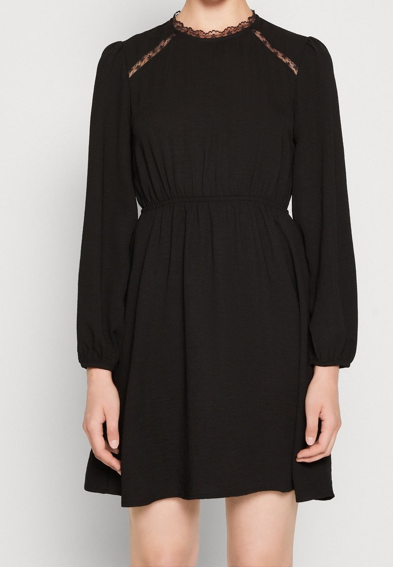 Mustat mekko, jossa on pitkät puff-hihat, pitsikoristeet hartioilla ja istuva vyötärö, valmistettu kevyestä kankaasta, jossa on pehmeä rakenne.