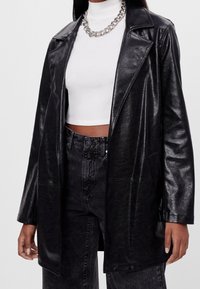 Czarna marynarka z faux skóry o błyszczącej fakturze, z klapami i dwoma przednimi kieszeniami. Noszona na białym swetrze z podwyższonym kołnierzem i dżinsami z wysokim stanem.