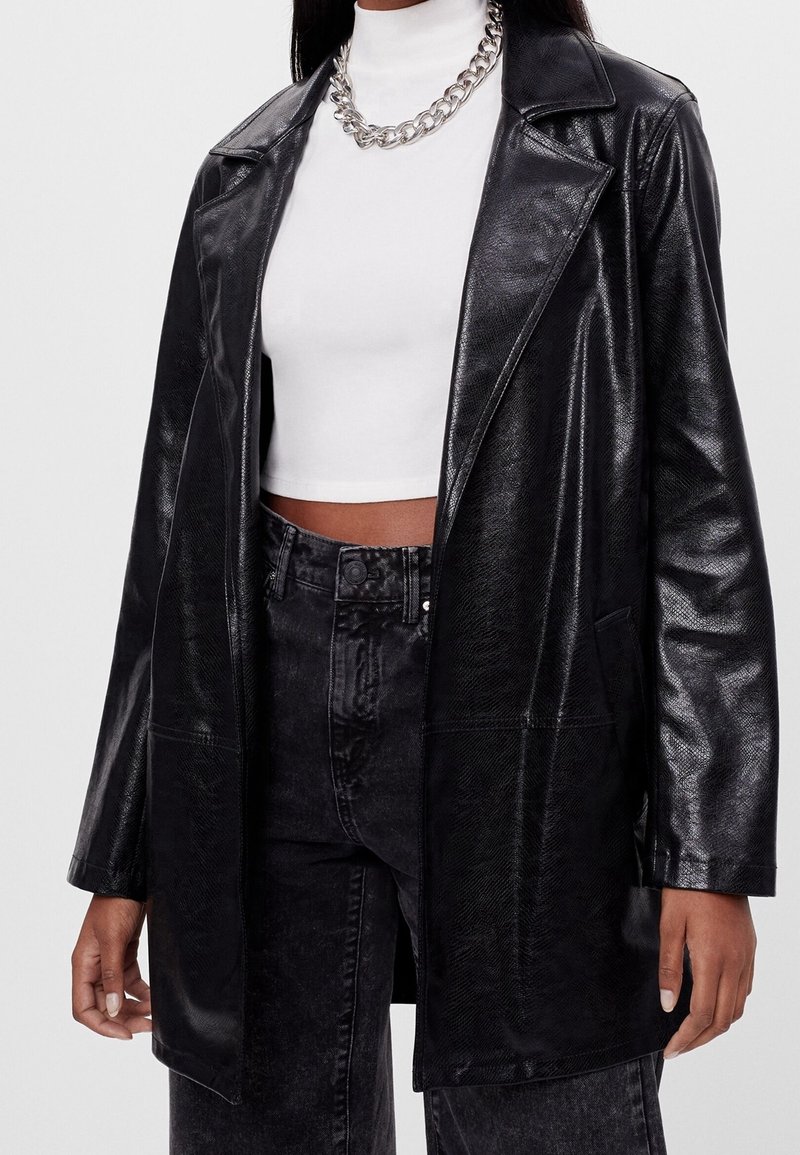 Czarna marynarka z faux skóry o błyszczącej fakturze, z klapami i dwoma przednimi kieszeniami. Noszona na białym swetrze z podwyższonym kołnierzem i dżinsami z wysokim stanem.