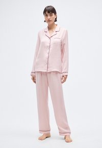 Ensemble de pyjama en soie rose comprenant une chemise boutonnée avec une poche et des accents de passepoil, ainsi qu'un pantalon ample assorti avec des détails à l'ourlet.