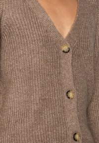 Cardigan marron tricoté avec un col en V, présentant trois grands boutons de couleur contrastante, mettant en valeur son tissu texturé.