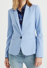 Ljusblå skräddarsydd blazer med slagkrage, enkel knappstängning och framfickor, i kombination med en mönstrad skjorta under.