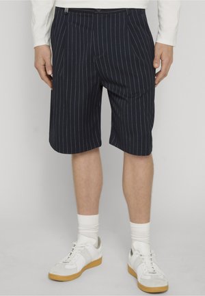 Marineblaue knielange Shorts mit Nadelstreifen, getragen mit weißen Socken und weißen Turnschuhen, kombiniert mit einem weißen langärmeligen Hemd.