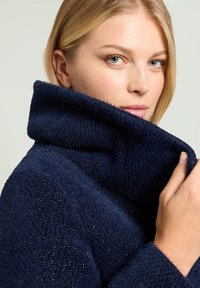 Blondynka o niebieskich oczach podnosi kołnierz ciemnoniebieskiego, teksturowanego płaszcza, patrząc lekko w bok na jednolitym tle.