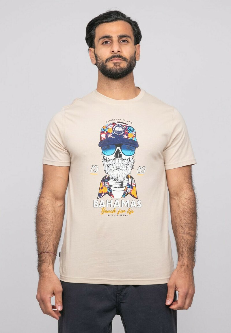 Homme portant un t-shirt beige avec un motif de crâne coloré en lunettes de soleil et chapeau, texte "Bahamas Beach for Life", et un pantalon foncé.