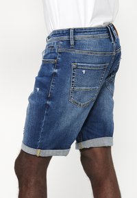 Kratke jeans hlače v temno modri barvi, z zavihkom na robu, uničenimi detajli in petimi žepi, opremljene z usnjenim logotipom na hrbtu.