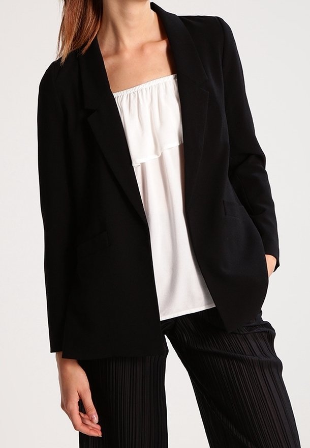 Blazer noir avec deux poches avant, revers structurés et coupe moderne, superposé sur un chemisier blanc et associé à un pantalon noir plissé.