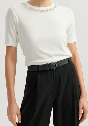 Femme portant un haut blanc à manches courtes avec un détail de collier en perles et un pantalon noir taille haute avec une ceinture noire à boucle argentée.