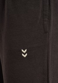 Pantalons de survêtement noirs en tissu doux, avec une finition texturée, des poches latérales et un logo en chevron blanc sur la jambe inférieure.